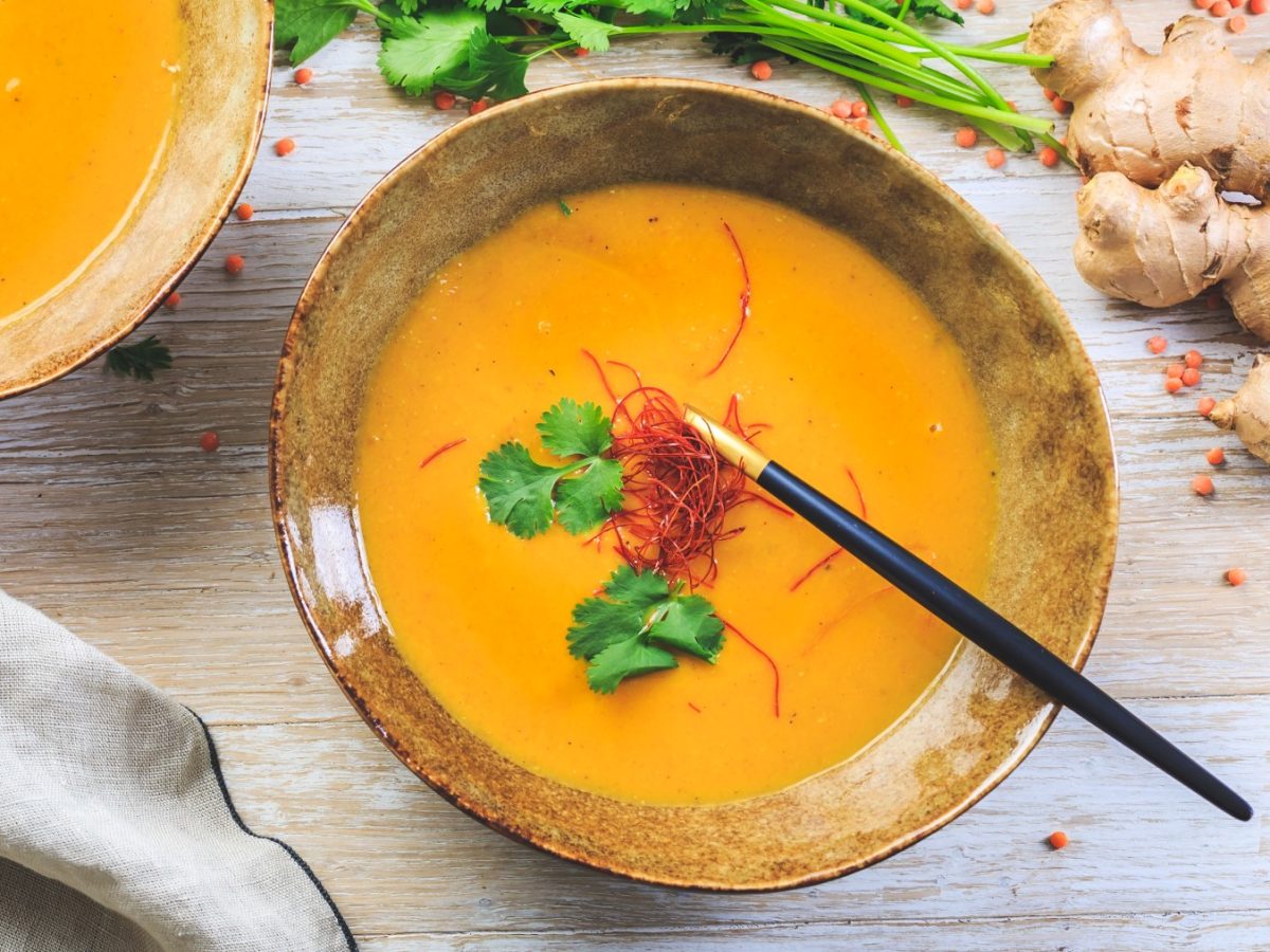 Miso-Suppe mit Ingwer und Karotten in einer Bowl. Rechts auf dem Bild Ingwer und Koriander, links unten ein Geschirrtuch. Der Hintergrund ist hellbraun.