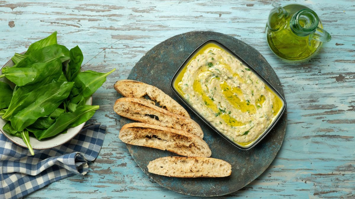 Draufsicht auf Zucchini-Dip auf einem teller mit Brot. Daneben frischer Bärlauch und Olivenöl in einer Karaffe.