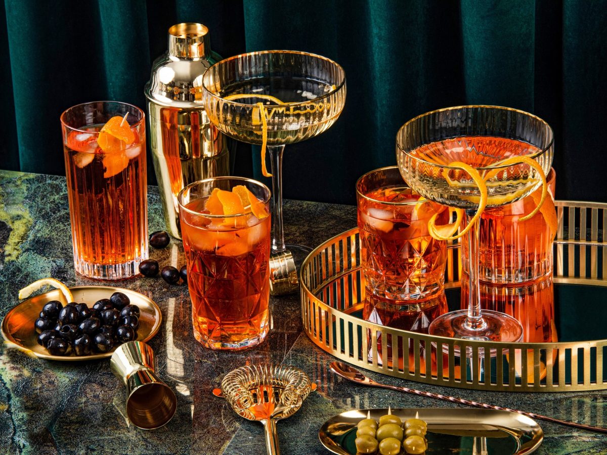 Mehrere Gläser, Cocktailzubehör und einige fertige Aperol Spritz Cocktails auf einem dunklen Marmortisch von schräg oben fotografiert.