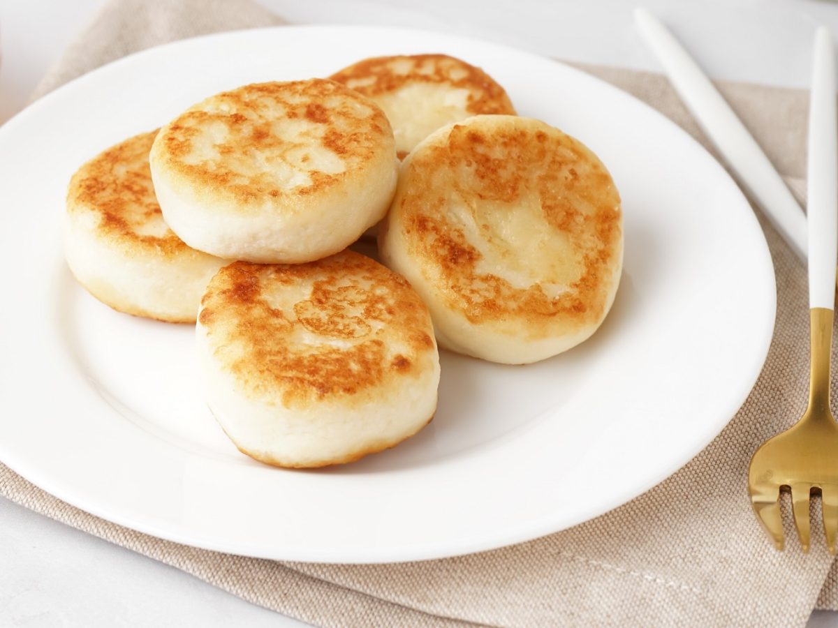 Fünf Pancakes mit Hüttenkäse auf einem weißen Teller und beigem Tuch. Alles von schräg oben auf hellen Untergrund fotografiert.