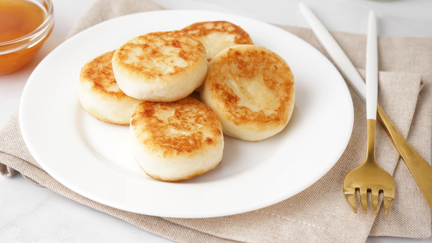 Fünf Pancakes mit Hüttenkäse auf einem weißen Teller und beigem Tuch. Alles von schräg oben auf hellen Untergrund fotografiert.