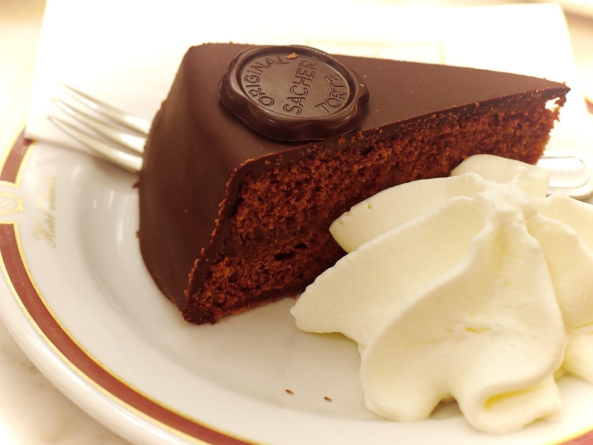Draufsicht schräg: Ein weißer Teller mit mit einem dünnen roten Rand. Auf dem ist ein Stück Sacher-Torte mit Sahne. Daneben liegt eine Kuchengabel.