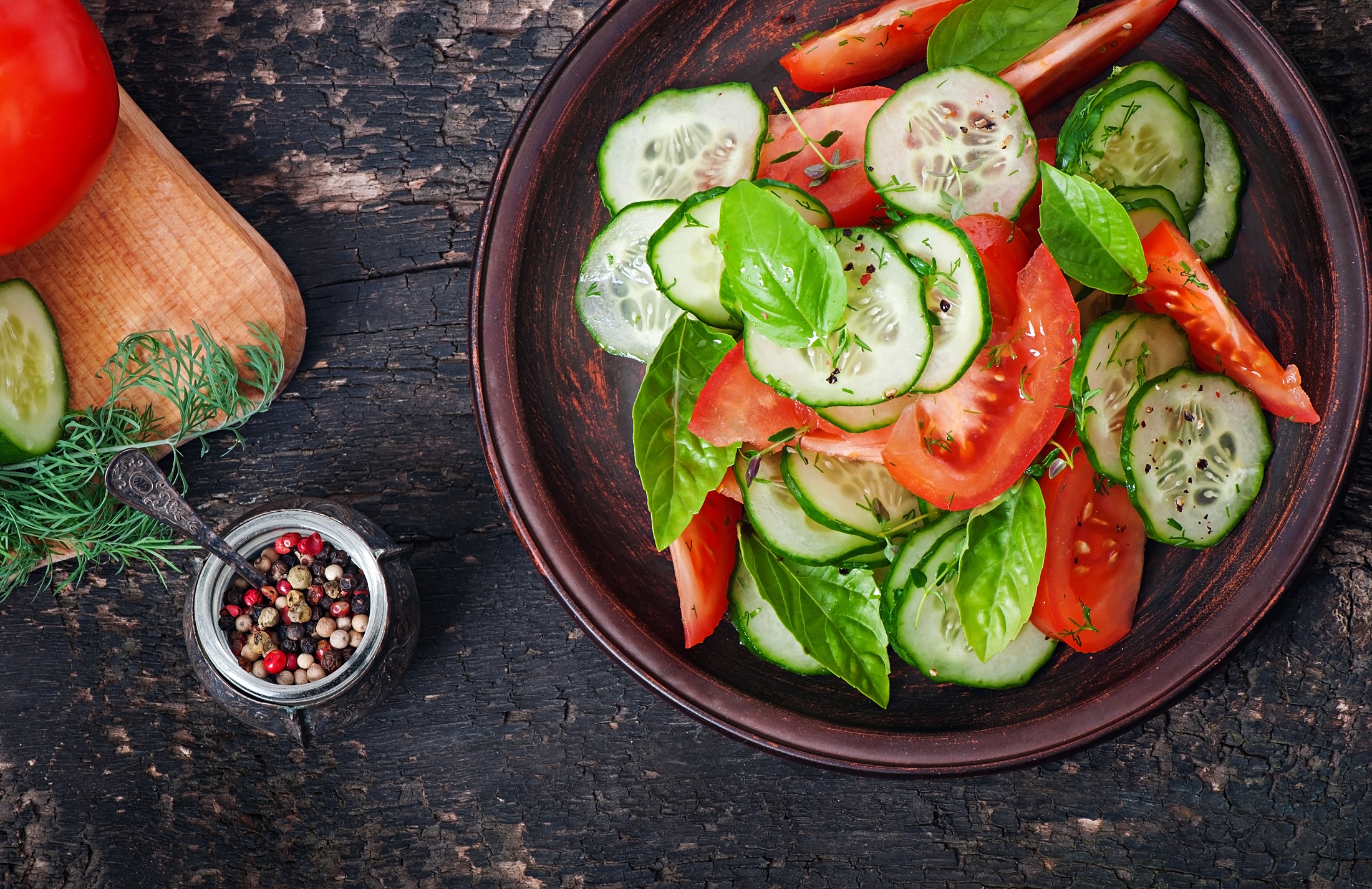 Draufsicht: Dunkler Untergrund mit einem Teller Tomaten-Gurken-Salat mit Basilikum. Daneben einen kleine Schale mit bunten Pfefferkörner und frischen Kräutern.