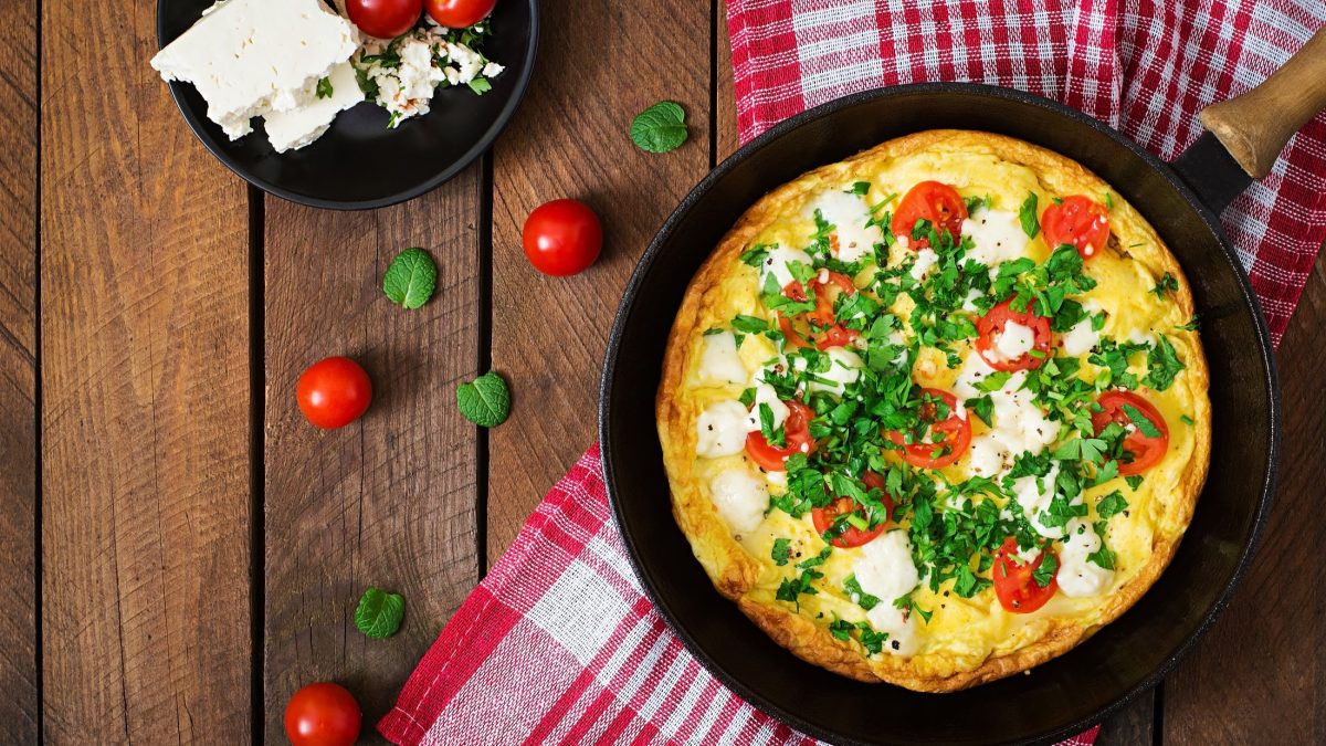 Draufsicht: Eine Pfanne mit einem Tomaten-Omelett mit Feta und Petersilie liegt auf einem rustikalen Holzuntergrund. Drum herum liegen frische Tomaten sowie ein rot-weiß-kariertes Küchentuch.