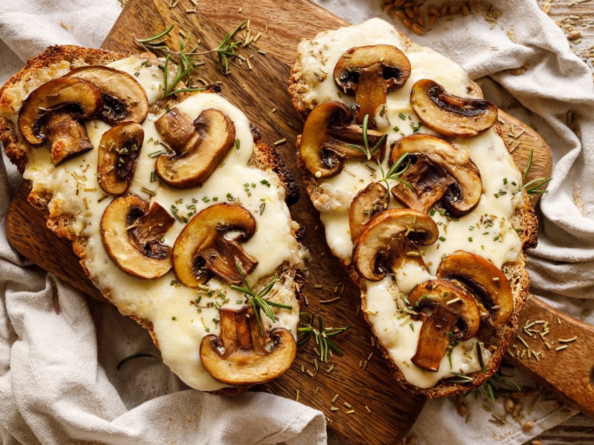 Draufsicht: Ein überbackenes Champignon-Käse-Sandwich liegt auf einem Holztisch. Daneben liegt ein überbackenes Käse-Sandwich mit Pilzen.