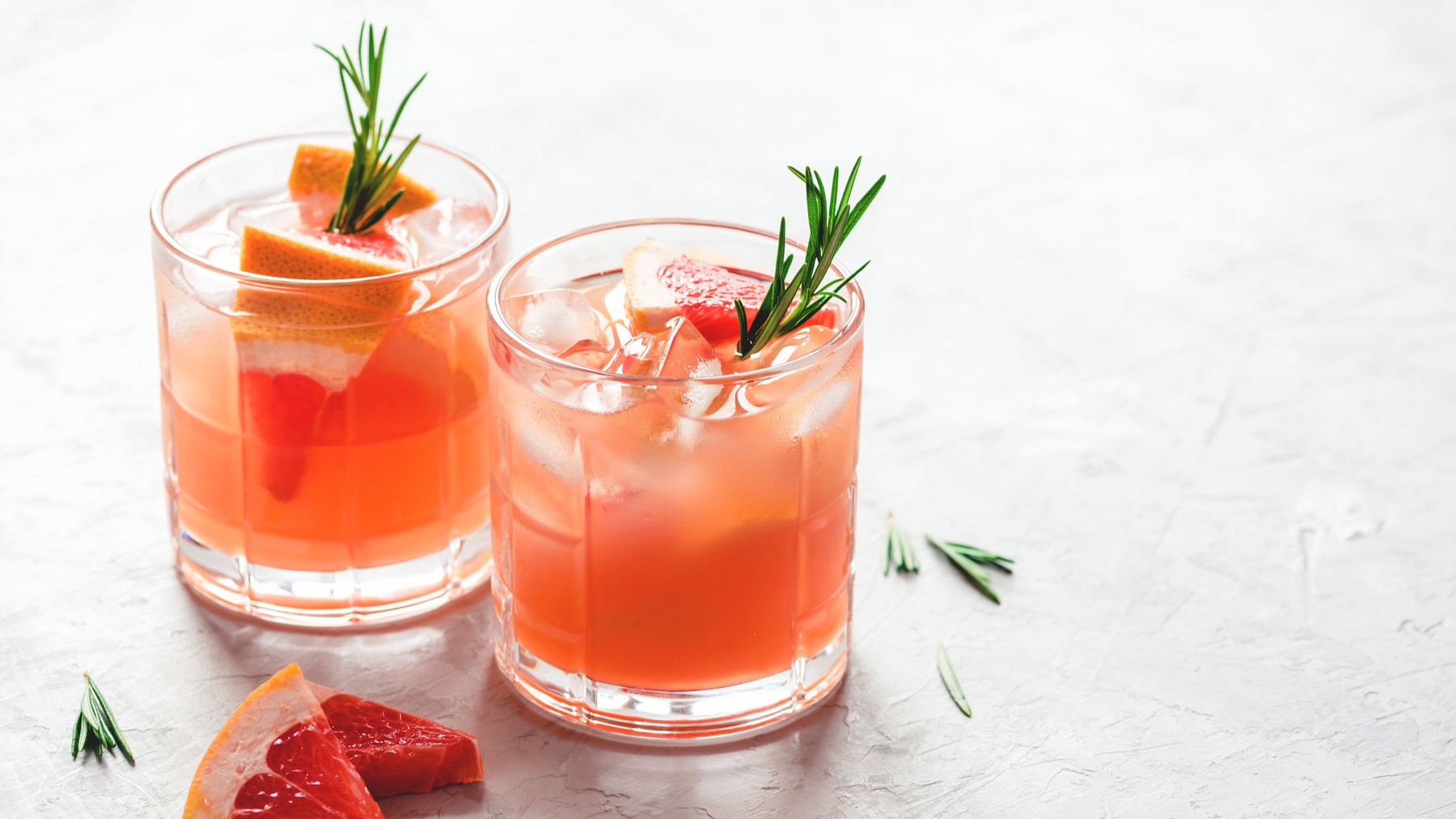 Zwei Gläser mit dem rötlichen Getränk auf einer weißen Tischfläche mit frischer Grapefruit und einigen Rosmarinnadeln. Alles leicht oben von der Seite fotografiert.