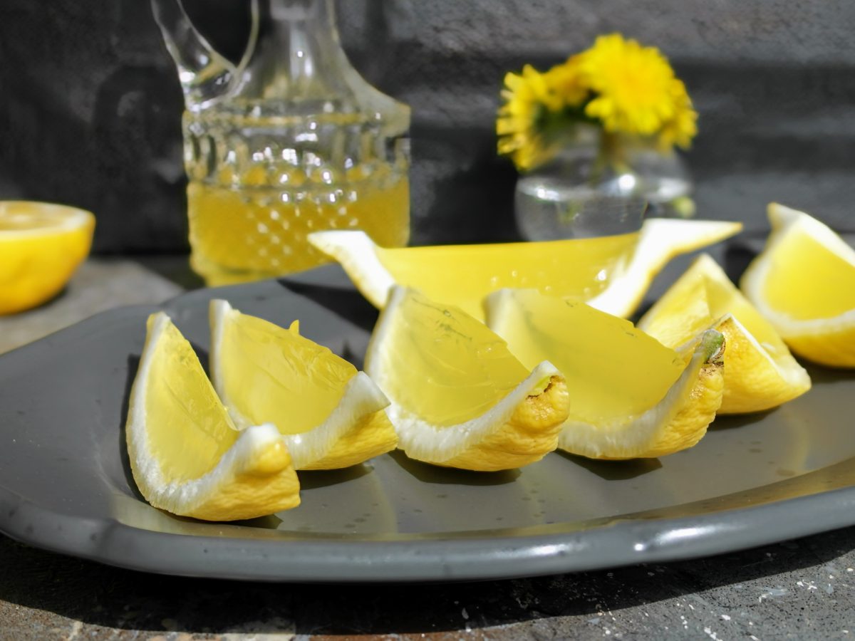 Mehrere Zitronenecken mit dem Limoncello-Gelee gefüllt auf einem schwarzen langen Teller und grauem Untergrund. Im Hintergrund eine Flasche mit Limoncello und einer halbierten Zitrone.