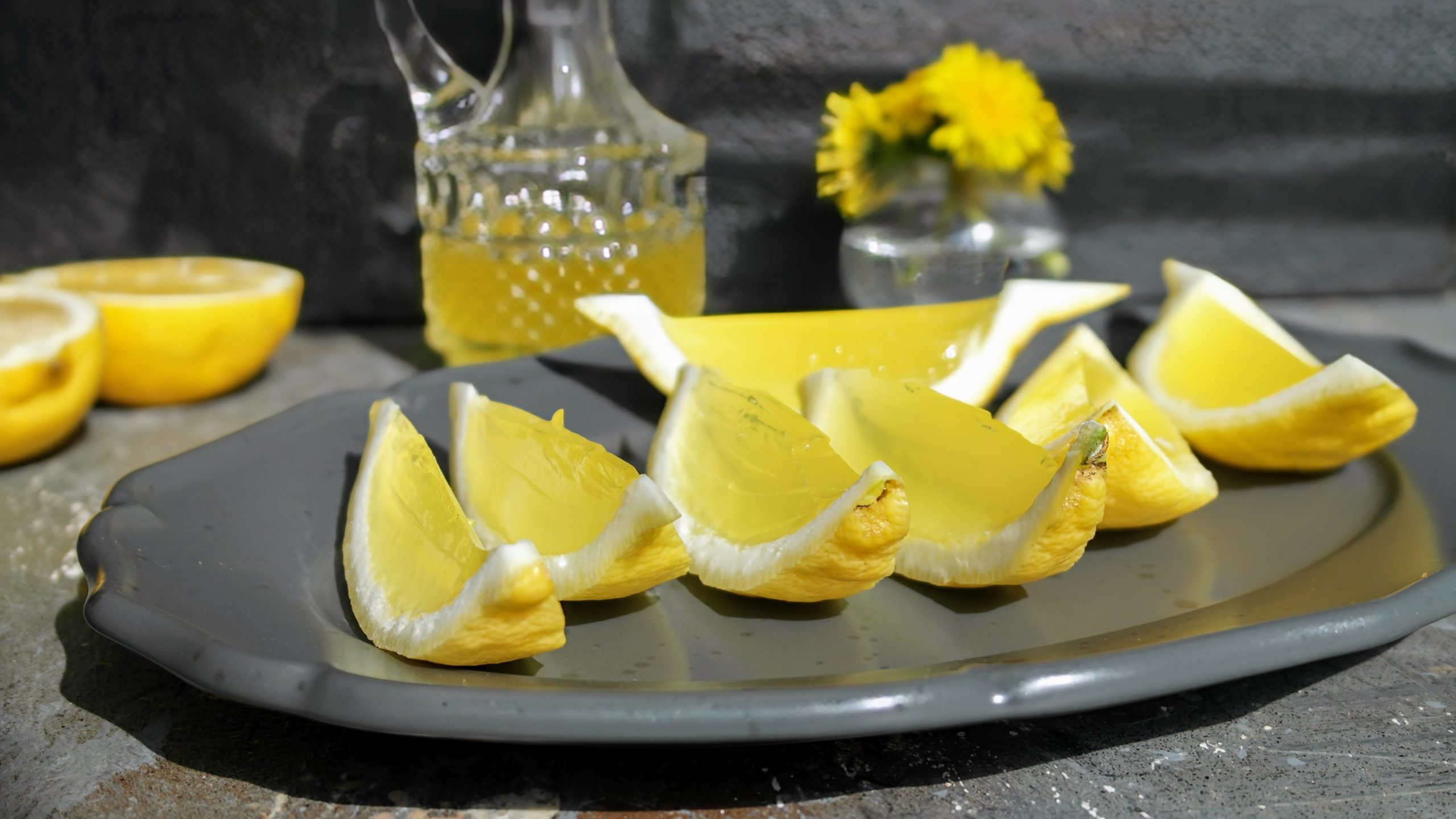Mehrere Zitronenecken mit dem Limoncello-Gelee gefüllt auf einem schwarzen langen Teller und grauem Untergrund. Im Hintergrund eine Flasche mit Limoncello und einer halbierten Zitrone.
