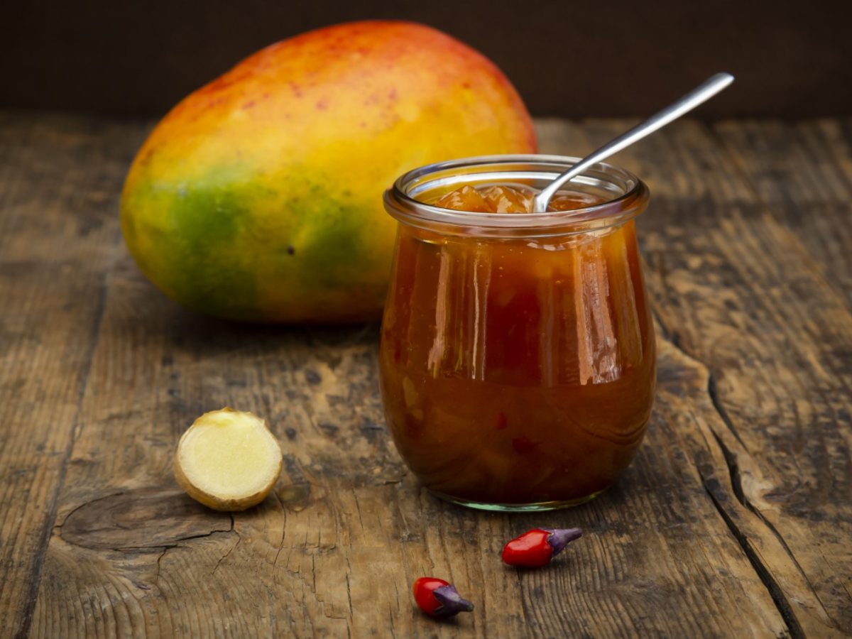 Mango-Chili-Chutney in einem Glas auf braunem Hintergrund. Hinten auf dem Foto eine Avocado.