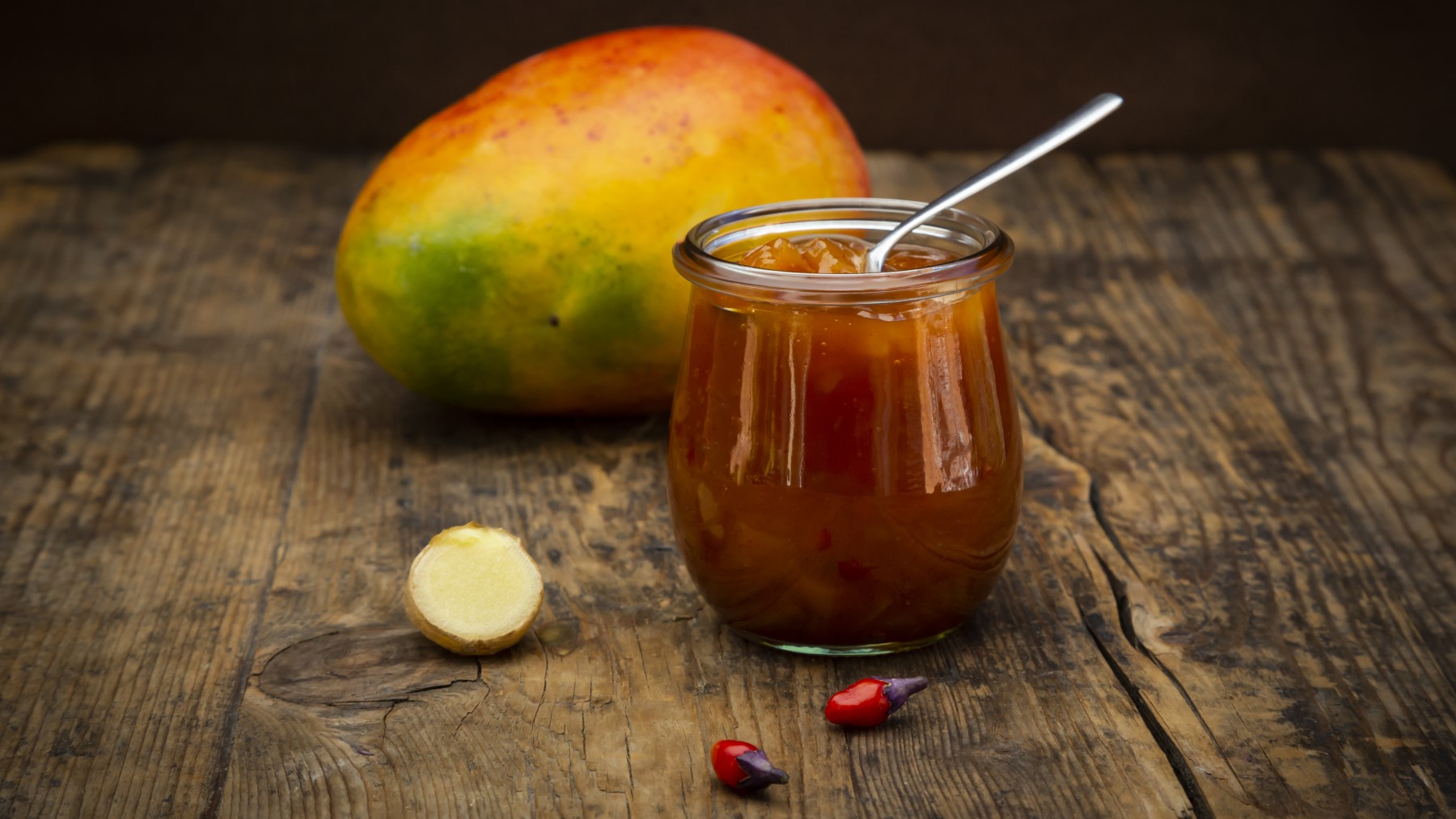 Mango-Chili-Chutney in einem Glas auf braunem Hintergrund. Hinten auf dem Foto eine Avocado.