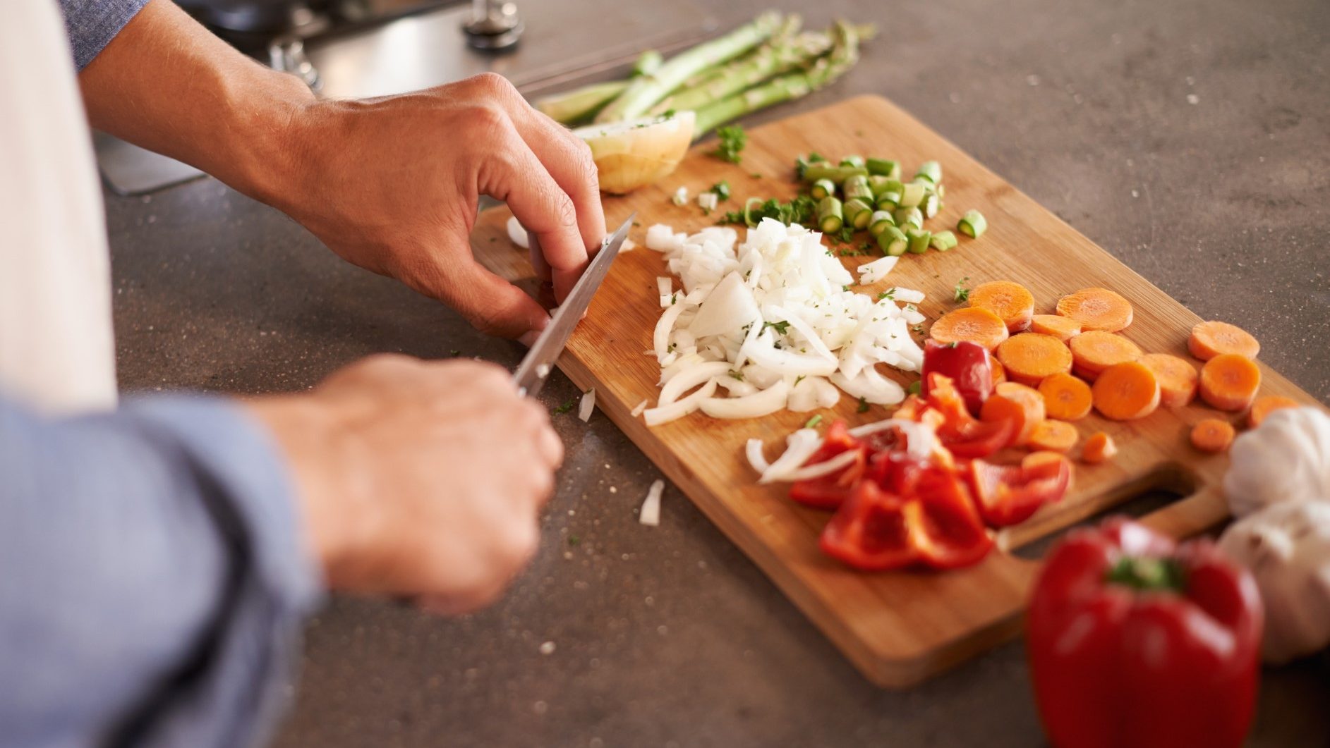 Hände zerteilen Zwiebeln, rohen Spargel und rote Paprika für gesunde Mahlzeit auf dunkler Arbeitsplatte.