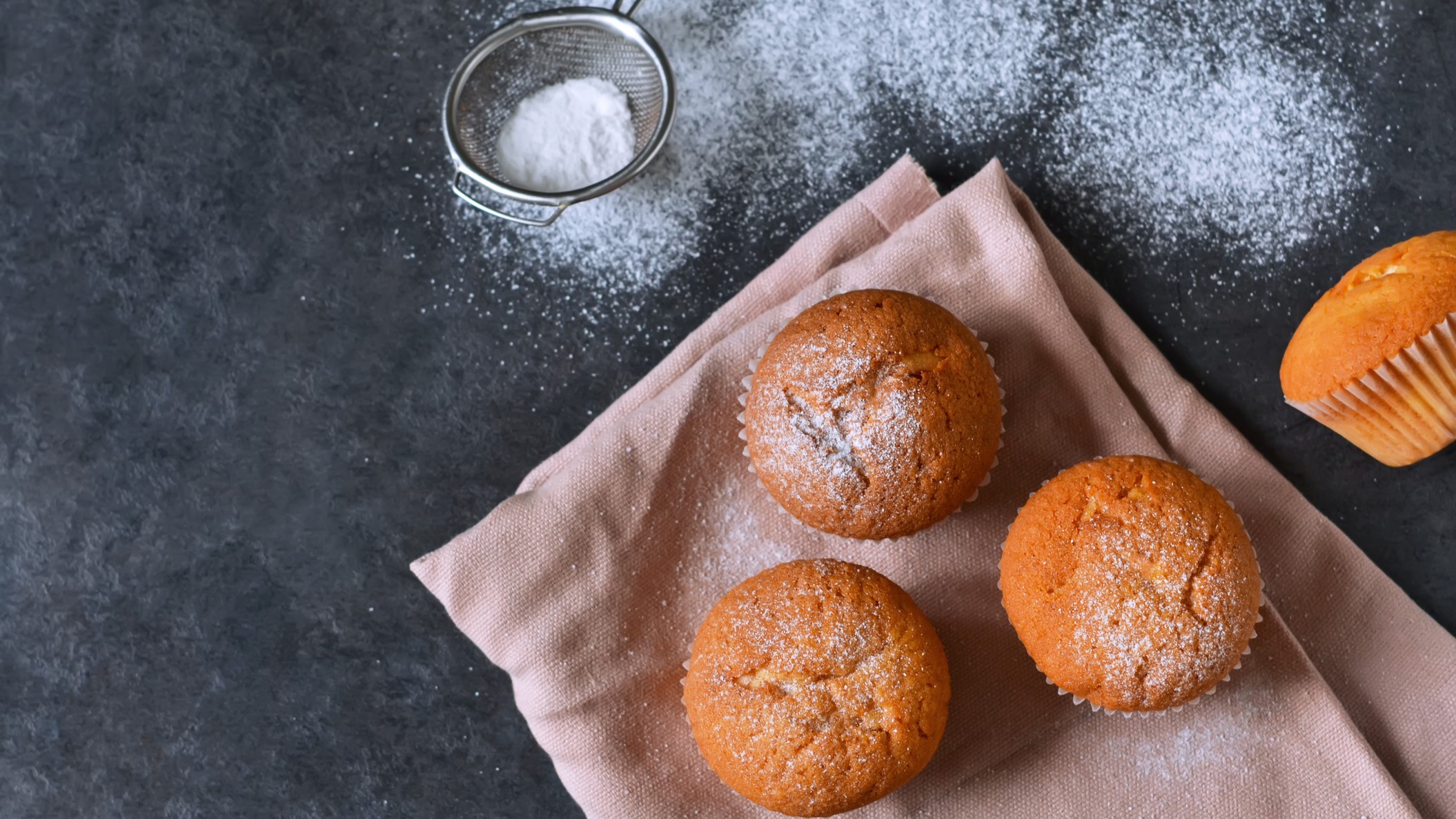 Vegane Muffins, mit Puderzucker bestäubt auf einem Küchentuch vor grauem Untergrund.