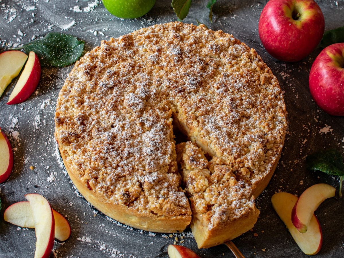 Angeschnittener Apfel-Streuselkuchen von oben fotografiert, daneben frische Äpfel.